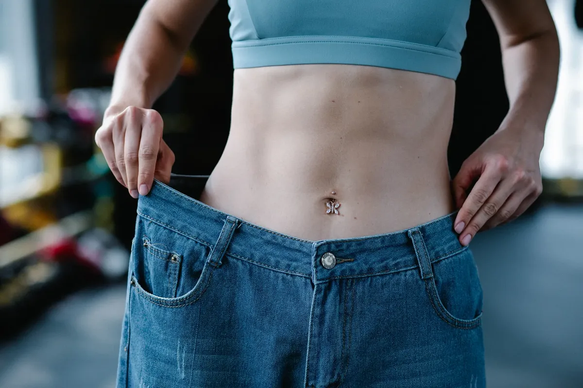 A woman shows her weight loss by holding oversized jeans revealing her toned stomach.