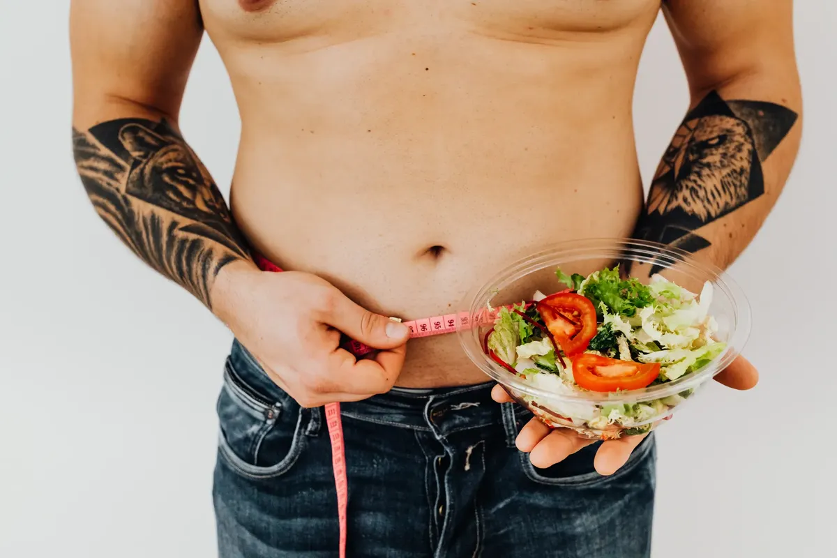 Tattooed man measuring waist while holding fresh salad, symbolizing healthy lifestyle and weight management.