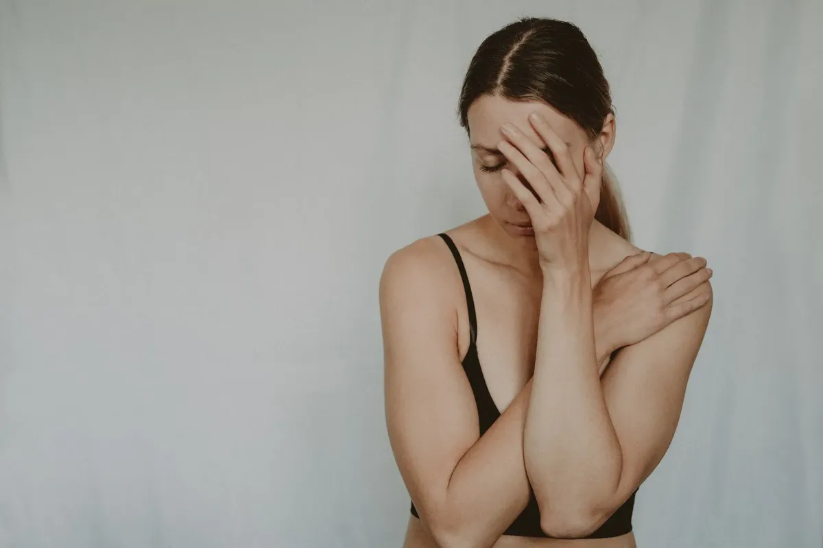 Young slender female with closed eyes covering breast and face on grey background